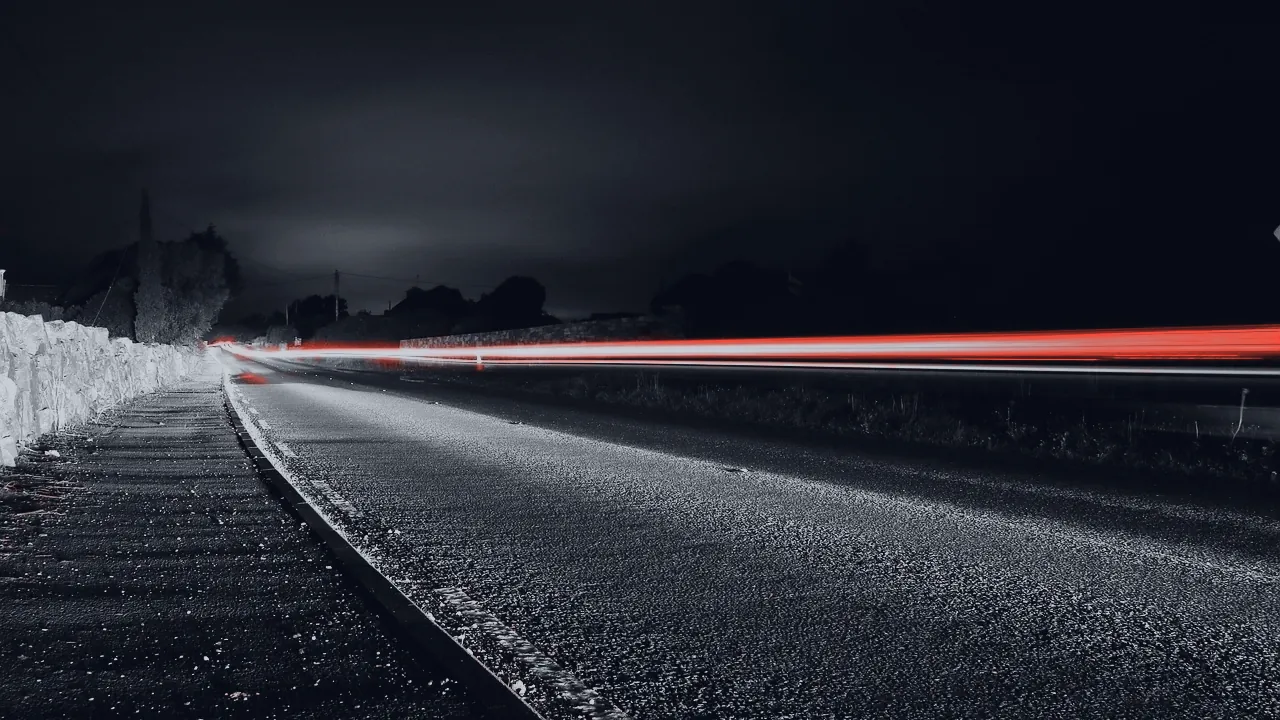 Une route de nuit, rayée par la traînée rouge des feux d’une voiture.